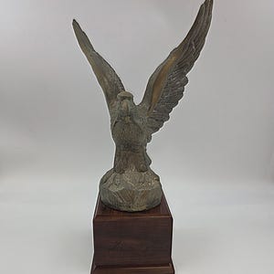 Puede incluir: Una escultura de águila de color bronce con las alas extendidas, montada sobre una base de madera oscura. Las alas del águila están detalladas con texturas de plumas, y la escultura tiene un aspecto envejecido.