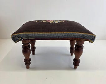 May include: A small, rectangular footstool with a dark brown, embroidered top featuring a floral design in pink, yellow, and green. The edges are trimmed with a striped pattern. It has four turned wooden legs, adding a touch of classic style.