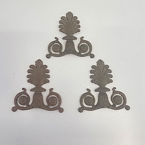 May include: Three antique bronze decorative metal ornaments. Each ornament features a central stylized leaf design atop a pedestal, with floral scrollwork on either side. The ornaments are arranged on a white surface.