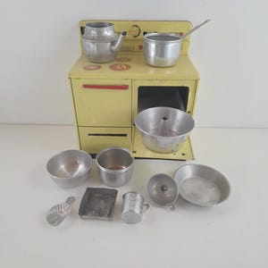May include: A vintage toy kitchen set featuring a yellow stove with a red handle and a collection of aluminum cookware. The set includes a teapot, saucepan, cake pan, bowls, and measuring cups, perfect for imaginative play.