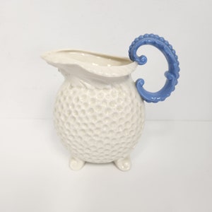 May include: A white ceramic pitcher with a blue handle. The pitcher has a textured surface and three feet.