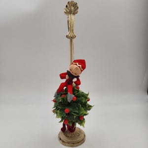 May include: A vintage Christmas decoration featuring a red felt elf doll sitting on a green holly sprig. The elf is wearing a red hat with gold trim and has a painted face. The holly sprig is attached to a white metal stand with a decorative top.