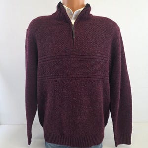 Può includere: Un maglione color bordeaux scuro con mezza zip, caratterizzato da un motivo a maglia testurizzato con fasce orizzontali a coste sul petto. Il capo presenta un colletto alto e una camicia bianca visibile sotto lo scollo.