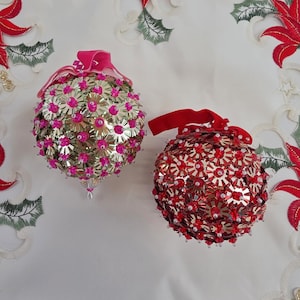 May include: Two spherical ornaments decorated with metallic flower-shaped sequins and small beads. One ornament features gold sequins with bright pink beads and a pink ribbon, while the other has red sequins with red beads and a red ribbon.