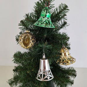May include: A small artificial Christmas tree decorated with four ornaments. The ornaments include a green bell, a gold sphere, a silver bell, and a gold bell-shaped ornament. The tree is set against a white background.