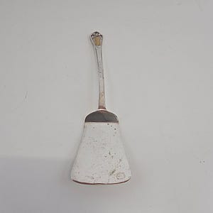 May include: A silver-toned metal serving utensil with a long, slender handle and a wide, flat, trapezoidal-shaped blade. The surface of the blade shows signs of wear with small dark spots and a circular hallmark near the bottom edge.