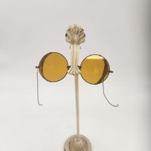 May include: Antique round yellow-tinted eyeglasses on a decorative stand. The glasses have a metal frame and a wire that loops down. The stand is off-white with an ornate floral design at the top and base.