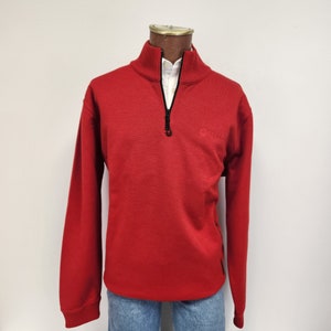 May include: A red long-sleeved sweater with a black zipper and a small logo on the right side. The sweater has a mock neck and a ribbed hem.