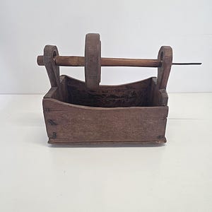 Può includere: Una scatola in legno marrone con base rettangolare e pannelli laterali curvi. Due supporti verticali sostengono un'asta orizzontale in legno che sporge sul lato destro. Una ruota circolare in legno è montata sull'asta sopra la scatola.