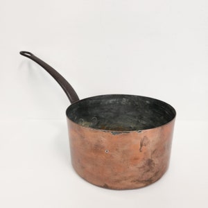 May include: A vintage copper saucepan with a dark brown iron handle. The saucepan has a worn, aged patina and is slightly dented.