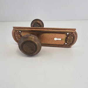 May include: Antique bronze door hardware set, featuring two round doorknobs and a rectangular backplate with a keyhole. The backplate has decorative floral accents on each end. The hardware has a vintage, aged appearance.