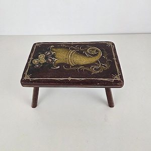 Puede incluir: Un pequeño taburete rectangular de madera con una superficie marrón oscuro. La parte superior está decorada con un diseño de cornucopia dorado y amarillo y detalles florales. El taburete tiene tres patas cortas de color marrón oscuro.