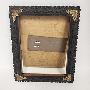 May include: Black wooden picture frame with ornate carvings and gold accents. The frame is empty and ready to display a photo or artwork.