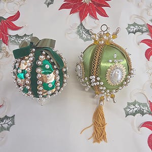 May include: Two spherical Christmas ornaments. The left ornament is dark green with gold metallic panels, white faux pearls, and a green velvet ribbon bow. The right ornament is light green satin with gold trim, white faux pearls, and a gold tassel.