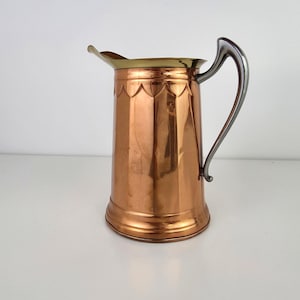 May include: A copper pitcher with a brass rim and a silver handle. The pitcher has a decorative scalloped design around the top. The copper has a warm, reflective finish, and the handle is curved.