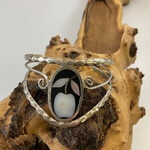 May include: Silver cuff bracelet with an oval black inlay featuring a white apple design with pink and white accents. The bracelet has a textured band and a twisted wire design, showcasing a unique fruit-themed jewelry piece.