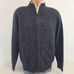 May include: A dark navy blue full-zip jumper featuring a textured knit pattern, a mock neck collar, and long sleeves. The garment has a central metal zip closure and ribbed cuffs and hem, worn over a light-coloured collared shirt.
