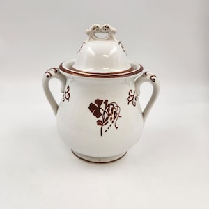Könnte beinhalten: Weißer Keramik-Zuckertopf mit Deckel und zwei Griffen. Die Schale hat braune Blumen- und Scroll-Designs. Der Deckel hat einen dekorativen Griff. Der Rand und die Akzente sind braun.
