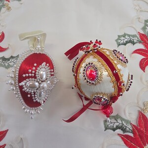May include: Two decorative ornaments. The left is a red, teardrop-shaped ornament adorned with white pearls and beads. The right is a spherical white ornament featuring red velvet ribbons, gold trim, iridescent cabochons, and dark red bead accents.