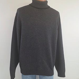 Könnte beinhalten: Ein dunkel anthrazitfarbener Langarmpullover mit geripptem Rollkragen, gerippten Bündchen und einem gerippten Saum. Das Strickkleidungsstück ist einfarbig gehalten und wird auf einem Schaufensterpuppen-Torso mit blauen Jeans präsentiert.