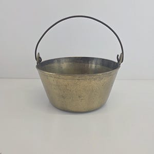 May include: A small, antique brass bucket with a dark metal handle. The bucket has a rounded base and a slightly flared rim. The brass has a warm, aged patina, suggesting it is vintage. The handle arches over the top, completing the classic design.
