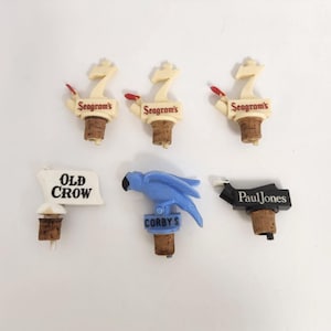May include: A collection of vintage bottle stoppers. The stoppers feature various designs, including a blue parrot, a sign that says "Old Crow", and several with the "Seagram's" logo. The stoppers are made of cork and plastic.