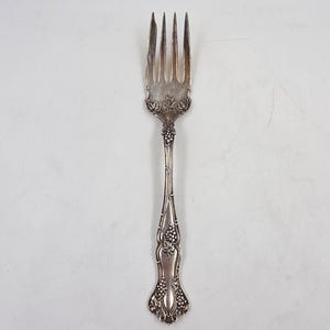 May include: A silver fork with four tines and ornate detailing. The handle features a grape cluster design and floral accents. The fork is likely vintage or antique, and is suitable for formal dining.