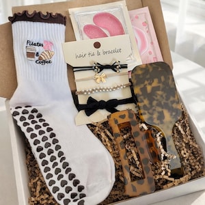 May include: A gift box containing a white sock with Pilates & Coffee embroidery, hair ties and bracelets, eye patches, a hairbrush, and hair clips. The sock has a brown ruffle and heart-shaped grips on the sole.