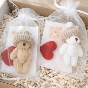 May include: Two small teddy bears, one tan and one white, each wearing a knitted hat with a pom-pom. They are packaged in clear bags with red heart accents and a small bear sticker, inside a cardboard box.