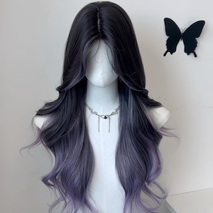 Op de afbeelding: Een lange, golvende pruik met een ombre-effect van donker naar lichtpaars. De pruik is gestyled op een mannequinhoofd, met een zilveren ketting. Een zwarte vlinderdecoratie staat op de muur achter de pruik.