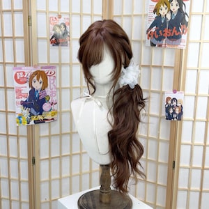 以下が含まれることがあります： 茶色のウィッグ。前髪とサイドポニーテールがあり、白い花のヘアアクセサリーで留められています。マネキンの頭に展示されています。背景には日本のアニメポスターがあります。