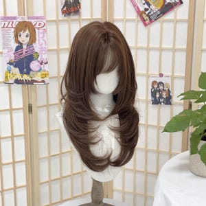 以下が含まれることがあります： 茶色のウィッグ。肩までのレイヤーヘアと前髪があります。マネキンの頭に展示されており、明るい背景です。ウィッグは自然なスタイルで、コスプレやファッション用です。