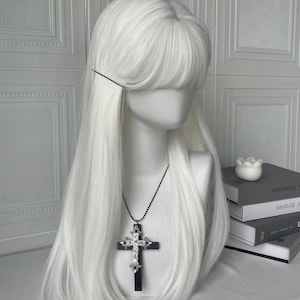 以下が含まれることがあります： 長いストレートヘアと前髪の白いウィッグがマネキンの頭にセットされています。シルバーのアクセントが付いた黒い十字架ペンダントネックレスがウィッグの前にぶら下がっています。背景には本と装飾的な要素があります。