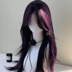 Puede incluir: Una peluca larga y en capas con reflejos morados oscuros y magenta. La peluca está peinada con flequillo y se muestra en una cabeza de maniquí blanca. También se ven un collar de rosas negro y una vela negra.
