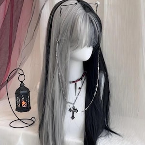 May include: A two-tone wig, split vertically with black and silver-grey strands, styled on a mannequin head. The wig features long, straight hair with a fringe. Accessories include a silver chain necklace with a cross pendant and a black choker.