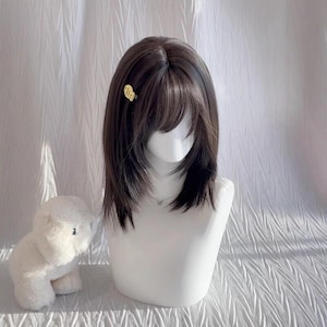 以下が含まれることがあります： 白いマネキンの頭に、レイヤーの入った毛先とサイドに流れる前髪のダークブラウンのウィッグが展示されています。小さな金のヘアクリップが横に留められています。白いぬいぐるみは左下の隅にあります。