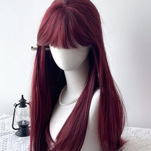 以下が含まれることがあります： マネキンの頭に展示された、長いストレートヘアと前髪のバーガンディのウィッグ。ウィッグは自然なヘアラインとソフトな仕上がりです。マネキンの首にはパールネックレスがあります。