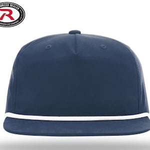 May include: A navy blue flat-brimmed cap with a white rope detail along the brim. The cap has a structured crown and a small button on top. The Richardson Cap Company logo is visible in the upper left corner.