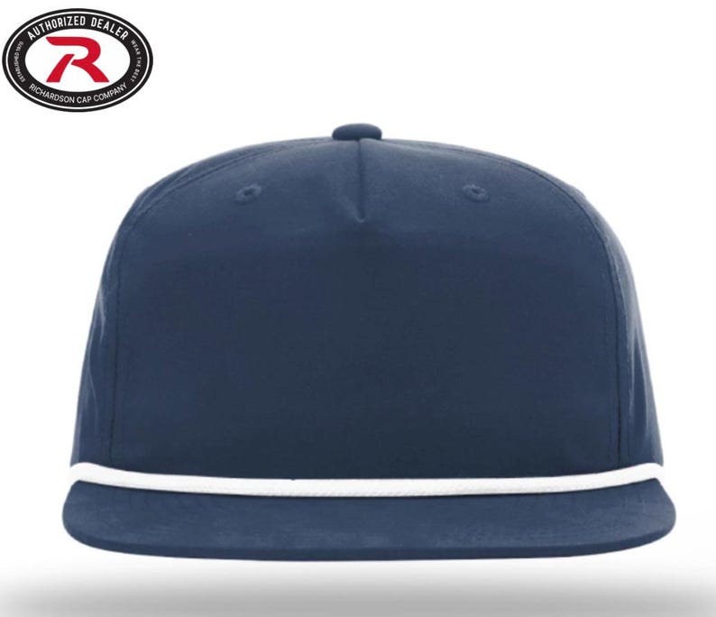 May include: A navy blue flat-brimmed cap with a white rope detail along the brim. The cap has a structured crown and a small button on top. The Richardson Cap Company logo is visible in the upper left corner.