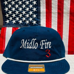 May include: A navy blue baseball cap with the text "Midlo Fire" in white script and the number "3" in red. The cap has a white rope detail and a flat brim. A tag on the brim reads "RICHARDSON".