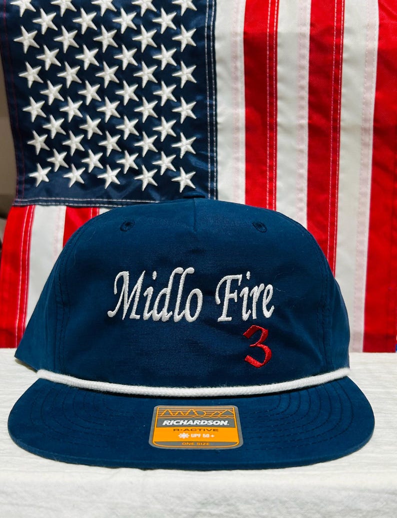 May include: A navy blue baseball cap with the text "Midlo Fire" in white script and the number "3" in red. The cap has a white rope detail and a flat brim. A tag on the brim reads "RICHARDSON".