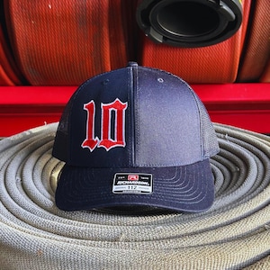 May include: A navy blue and black mesh trucker hat with a red and white embroidered number "10" on the front. The hat has a black adjustable strap with a white label that reads "Richardson. Est. 1970. Original Trucker. 112. Adjustable."
