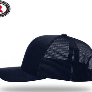 May include: A navy blue trucker hat with a curved brim and a mesh back. The hat features a structured front panel and a snapback closure. The Richardson Cap Company logo is visible on the top left.
