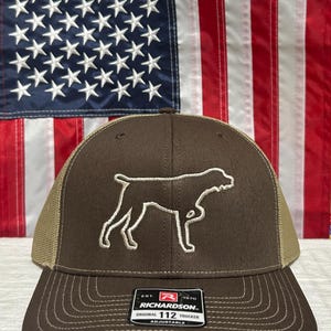 May include: A brown trucker hat with a tan mesh back and a white embroidered outline of a dog. The hat has a curved brim and the text "RICHARDSON ORIGINAL 112 TRUCKER" on a tag.