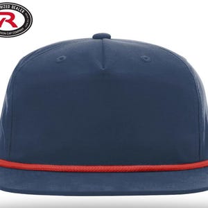 May include: A navy blue flat-brimmed cap with a red rope detail along the brim. The cap has a structured crown and a small button on top. The Richardson Cap Company logo is visible in the upper left corner.