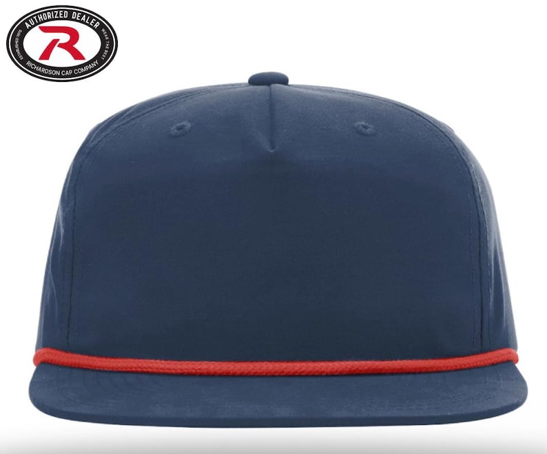 May include: A navy blue flat-brimmed cap with a red rope detail along the brim. The cap has a structured crown and a small button on top. The Richardson Cap Company logo is visible in the upper left corner.