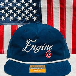 May include: A navy blue baseball cap with the word "Engine" embroidered in white script and the number "6" in red. The cap has a white rope detail and a flat brim. The Richardson brand tag is visible.