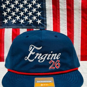 May include: Navy blue cap with a flat brim, featuring the word "Engine" in white script and the number "26" in red. A red rope detail accents the brim. The background includes an American flag.