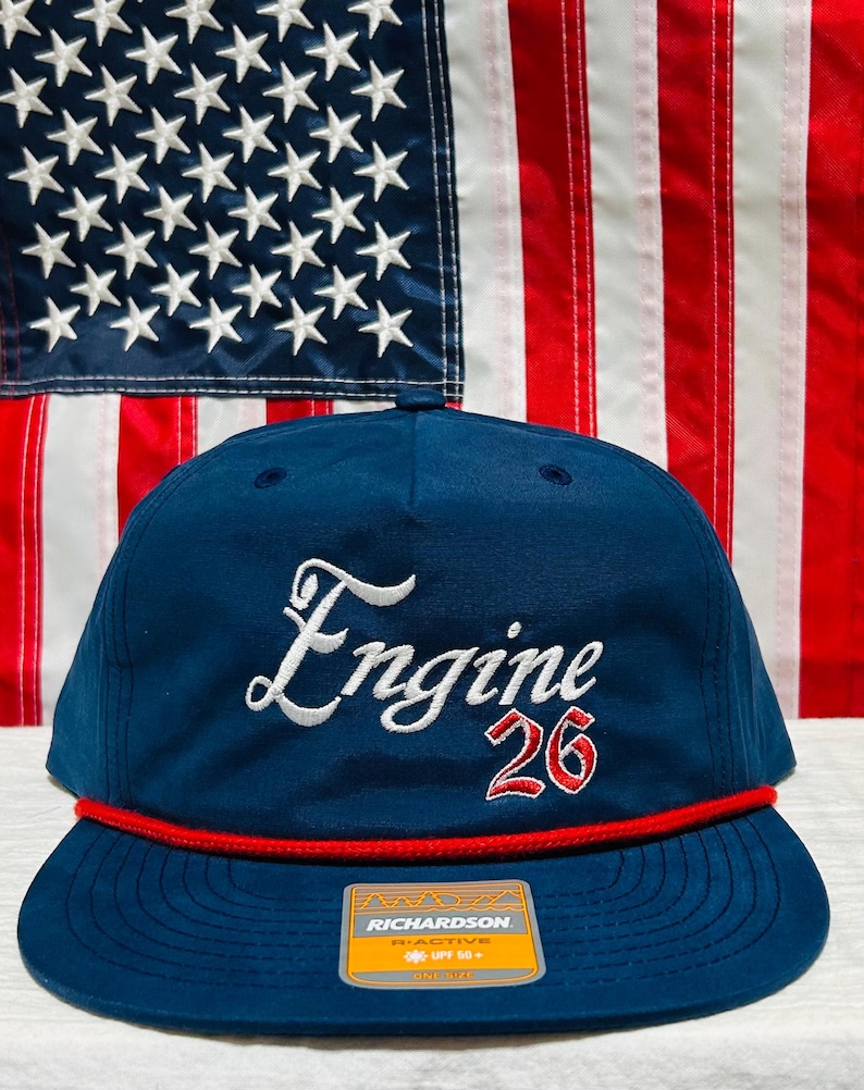May include: Navy blue cap with a flat brim, featuring the word "Engine" in white script and the number "26" in red. A red rope detail accents the brim. The background includes an American flag.
