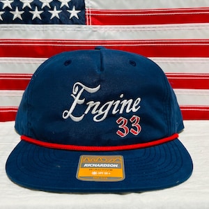May include: Navy blue cap with "Engine 33" embroidered in white and red. Features a red rope detail along the brim. The cap has a flat brim and a Richardson tag.